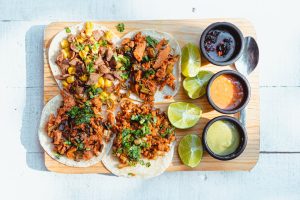 Colorful vegan tacos with lime slices and dipping sauces on a wooden tray, shot in bright light.